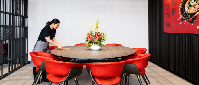 Dining area with a large round wooden table surrounded by eight red chairs. A floral arrangement sits at the center of the table. A person is standing to the left, adjusting something on the table. The background features black walls and a piece of artwork displaying Chinese characters and an image of food.