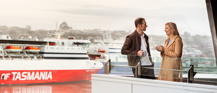 wo people standing on a balcony or terrace, engaged in conversation. In the background, a large ferry with the text 'SPIRIT OF TASMANIA' is visible, set near a harbor or port with other ships and buildings in the distance.