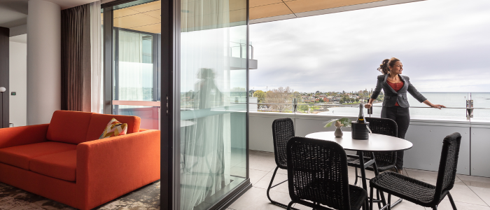 Modern indoor-outdoor living space with an orange sofa inside next to a sliding glass door. The door opens to a balcony with a round table and four black chairs. A person stands on the balcony, looking out over a view of buildings and trees under a cloudy sky.