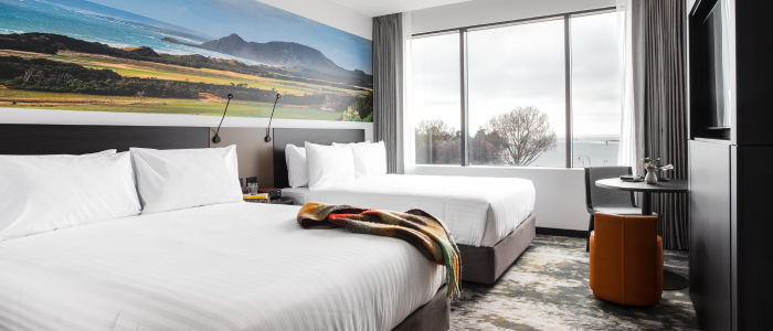 Modern hotel room with two neatly made double beds featuring white linens and pillows. A large mural above the headboards depicts a scenic landscape with mountains, greenery, and the ocean. The room includes a large window letting in natural light, with a view of trees and possibly water in the distance. A small round table with an orange stool is near the window, and wall-mounted reading lights are above the beds.