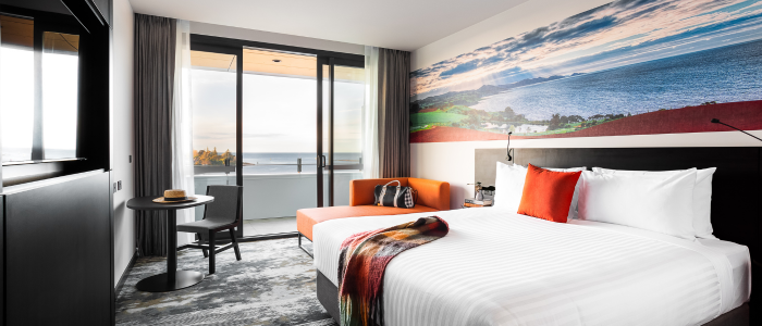 Modern hotel room with a large bed, an orange sofa, and a small round table with two chairs. A large window or sliding door opens to a balcony with an ocean view. The wall behind the bed features a mural of a coastal landscape. A blanket is draped over the bed, and decorative pillows are placed on the sofa.