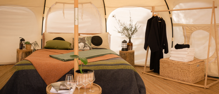 Interior of a cozy, well-decorated tent featuring a central bed with earthy-toned pillows and blankets. Wooden stump nightstands with lanterns and plants flank the bed. A small round table with wine glasses and a book sits in front, and a wooden clothing rack with clothes and towels stands beside a wicker basket to the right. Natural light filters through the tent’s fabric walls.