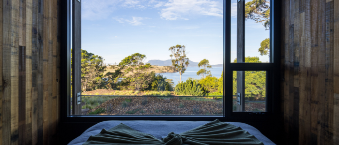 View from inside a room through a large window framing a scenic landscape with trees, greenery, and a body of water. The sky is partly cloudy. In the foreground, part of a bed with neatly arranged bedding is visible.