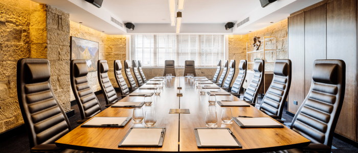 A modern conference room with a long wooden table surrounded by twelve high-back leather chairs. Each seat is set with a notepad and a glass of water. The room features stone walls, large windows with blinds, ceiling lights, mounted speakers, and a piece of artwork on one wall.