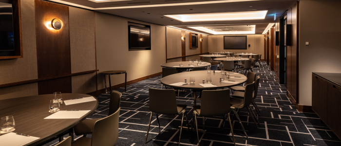 A modern conference room featuring round tables surrounded by chairs, each table set with glasses of water, notepads, and pens. The room has a patterned carpet, wood-paneled walls, recessed ceiling lighting, and several flat-screen televisions mounted on the walls.