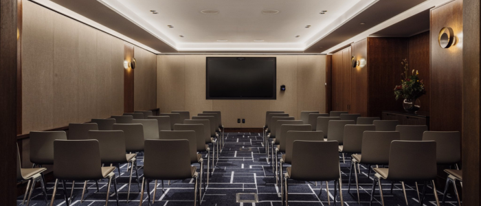 conference room with rows of gray chairs facing a large flat-screen TV mounted on the front wall. The room features beige walls, dark wood paneling, a patterned carpet, wall sconces for lighting, and a flower arrangement on a table to the right.