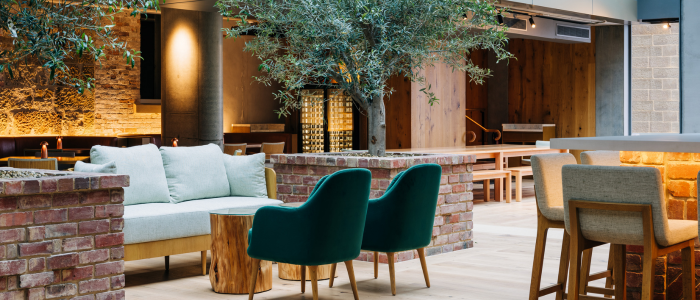 A modern indoor seating area with a light-colored sofa, two green chairs, and a wooden table in the foreground. The background includes brick walls, additional wooden tables and chairs, and an olive tree as part of the decor. Warm lighting enhances the cozy and inviting atmosphere.