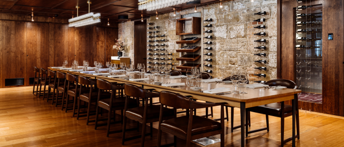 An elegant dining room featuring a long wooden table set for a meal with plates, glasses, and cutlery. Wooden chairs surround the table, and the room has wooden floors and walls. One wall is lined with wine racks filled with bottles, and modern light fixtures hang from the ceiling.