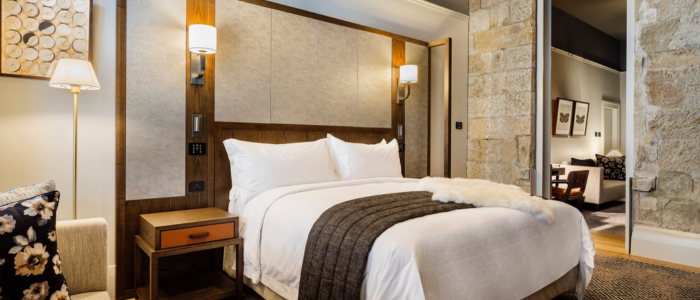 A modern, well-decorated bedroom featuring a large bed with white sheets, pillows, and a dark gray blanket. The bed is centered against a tall wooden headboard with two wall-mounted lamps. A small bedside table with a drawer and power outlet sits to the left. The room has light-colored walls and a stone accent wall on the right. An open doorway in the background reveals a seating area and framed pictures.