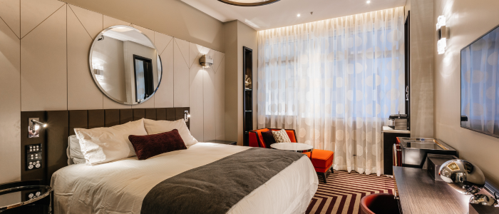 A stylish hotel room with a large bed dressed in white linens and a dark gray throw. A round mirror hangs above the bed, flanked by two bedside tables with lamps. The room includes a window with sheer curtains, two orange chairs, and a small round table. A flat-screen TV is mounted on the wall above a desk with an upholstered chair. The floor features a striped carpet in brown and beige tones.