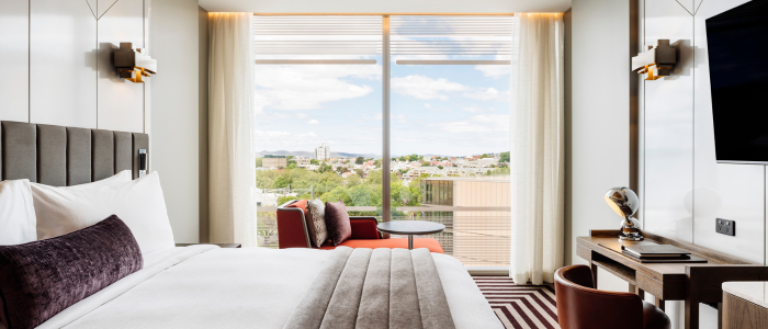 A modern hotel room with a large bed dressed in white linens, a dark gray blanket, and a dark red bolster pillow. A round mirror hangs above the bed, with wall-mounted reading lights and a bedside table holding an alarm clock and phone. An orange armchair with an ottoman sits by a window with sheer curtains. The room features striped brown and beige carpeting, and a flat-screen TV mounted above a desk area.