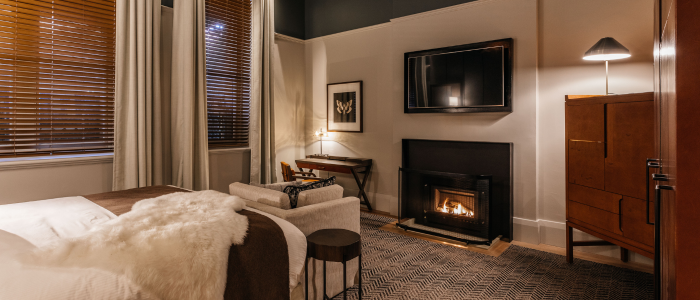 A cozy, modern bedroom featuring a bed with white and brown bedding, a small side table, and an armchair. A lit fireplace with a flat-screen TV mounted above it adds warmth to the room. To the right of the fireplace is a wooden cabinet with a lamp. A desk and chair sit in the corner beneath a piece of wall art. Large windows with wooden blinds and white curtains let in natural light.