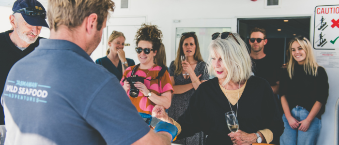 A group of people are gathered on the deck of a boat, a person wearing a shirt with 'Tasmanian Wild Seafood Adventures' on the back is interacting with another person holding a glass of wine. Several others are in the background, some with cameras or drinks.
