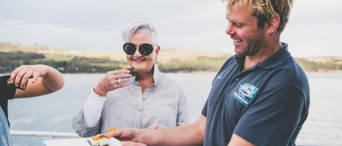 Three people are standing aboard a boat. The person on the right wears a dark blue shirt with a logo and is holding a tray of freshly caught seafood. The middle person, with short gray hair, wears a light-colored shirt. The person on the left is partially visible, holding an object near their mouth. The background features water and distant green-covered land.