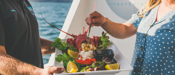 Two people hold a large white rectangular plate with a seafood dish featuring a lobster, lemon wedges, leafy greens. One person is using a fork to take some food.
