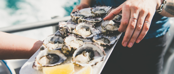 A close-up of a person holding a tray of opened oysters, arranged with lemon wedges. Another hand reaches toward the tray, taking an oyster.