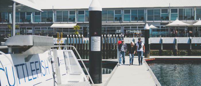 A dock with a boat named 'CUTTLEFISH' on the left side. Four people are walking on the dock toward the camera. In the background, a building with large windows and outdoor seating under white umbrellas is visible.