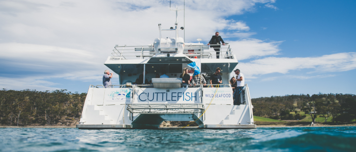A large white boat named 'Cuttlefish Wild Seafood' is docked on a body of water. Several people are standing on the deck engaging in conversation. The background features a scenic view of trees and hills under a partly cloudy sky.