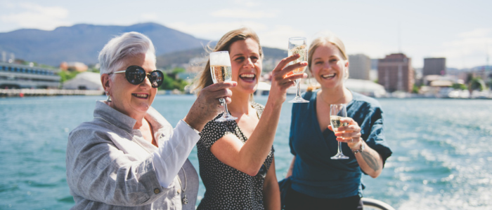 Three people stand on a boat, raising glasses in a toast. The background features a body of water, buildings, and mountains.