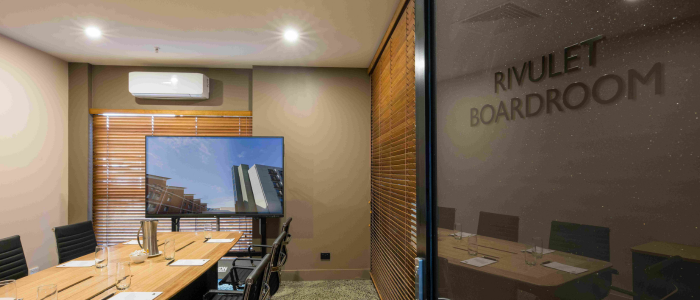 A modern boardroom with a long wooden table surrounded by black chairs. The table is set with notepads, pens, glasses, and a pitcher of water. A large screen at one end displays an image of buildings. The room features neutral-toned walls, wooden blinds on the windows, and an air conditioning unit above the screen. The glass door bears the text 'RIVULET BOARDROOM' in capital letters.