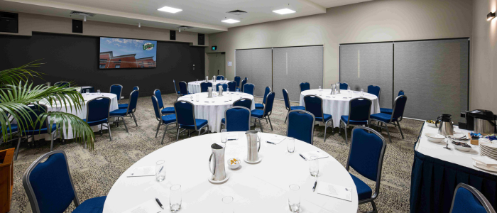 conference room with round tables covered in white tablecloths, each surrounded by blue chairs. The tables are set with water pitchers, glasses, notepads, and small bowls. The room features a carpeted floor, neutral-colored walls, and a large screen displaying an image of a building with the text 'Quest.' Plants are visible in the foreground, and a refreshment station with coffee pots and cups is on the right.