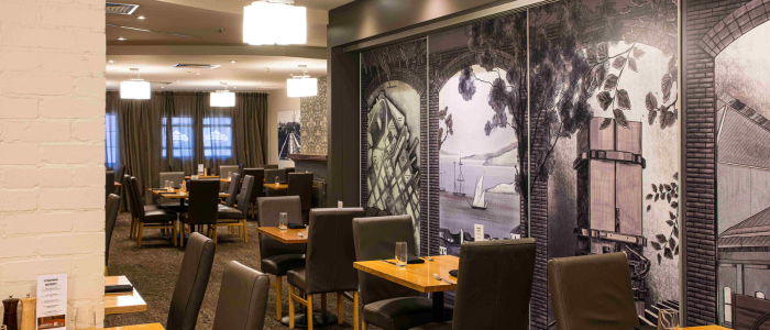 A dining area with several neatly arranged tables and chairs. The walls are adorned with large artistic murals featuring scenes such as a ship and architectural elements. Overhead lighting provides a calm and inviting ambiance.