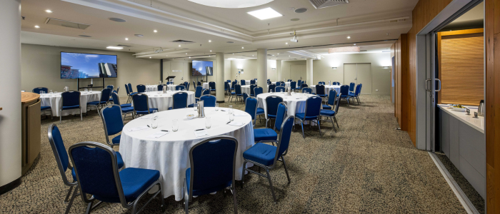 A conference or banquet room with multiple round tables covered in white tablecloths, each surrounded by blue chairs. The room has a carpeted floor, recessed ceiling lighting, and two large screens displaying an image of a building.