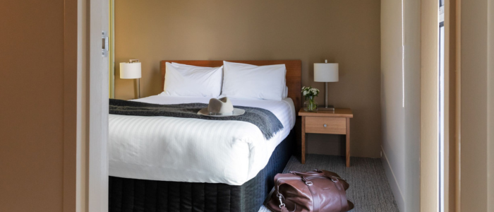 A neatly made bed with white linens and two pillows, set against a beige wall. Two identical bedside tables with lamps flank the bed, one also holding a small plant. A hat rests on the bed, and a brown leather bag is on the carpeted floor near the foot of the bed. Natural light brightens the room.