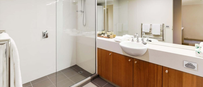 A modern bathroom featuring a glass-enclosed shower with a chrome showerhead and square floor drain on the left. To the right, a long countertop holds a white sink with a chrome faucet and neatly arranged toiletries. Wooden cabinets with silver handles sit below the counter. A towel rack and electrical outlet are visible on the wall in the background.