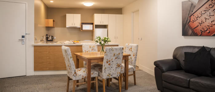A modern, well-lit kitchen and dining area with wooden lower cabinets and white upper cabinets. The kitchen includes a microwave, kettle, and small appliances on the countertop. A dining table with four floral-patterned chairs sits in the center, topped with a vase of flowers. To the right, a black leather sofa with cushions is positioned beneath wall art.