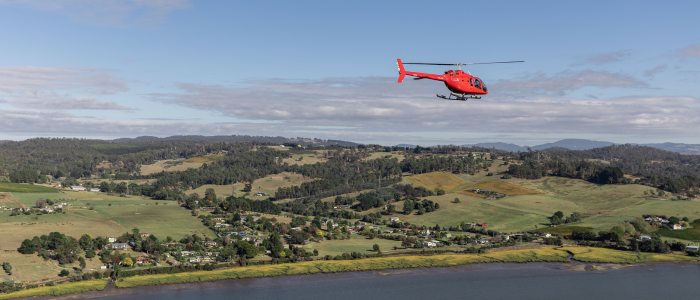 A red helicopter is flying over a scenic landscape featuring rolling green hills, scattered trees, and a river in the foreground. The sky is partly cloudy with patches of blue visible. In the distance, there are more hills and a mix of open fields and wooded areas.