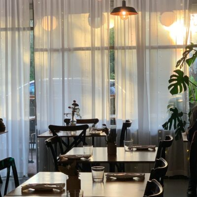 Interior of a Le Coq with neatly arranged tables and chairs, set with napkins and glasses. Three pendant lights hang from the ceiling, casting a warm glow. Large windows with sheer white curtains allow natural light to enter. A potted plant is visible on the right side, adding greenery to the space.