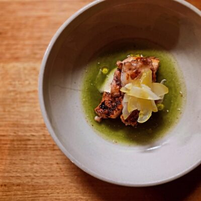 Dish featuring a piece of grilled or roasted meat, possibly fish or chicken, served in a shallow bowl with thinly sliced vegetable garnish and green sauce. The bowl rests on a wooden surface.