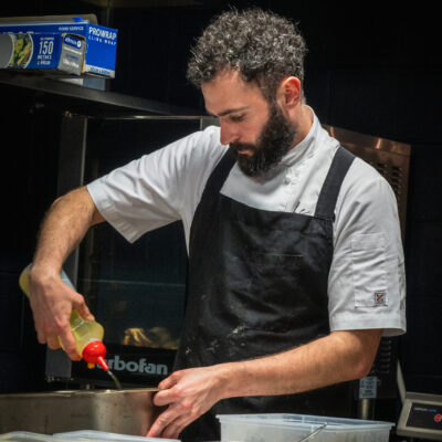 Chef Tristan Punelle in a kitchen wearing a white shirt and black apron, pouring liquid from a bottle into a container on the counter. Surrounding them are kitchen items like plastic containers, a metal pot, and green cloth.