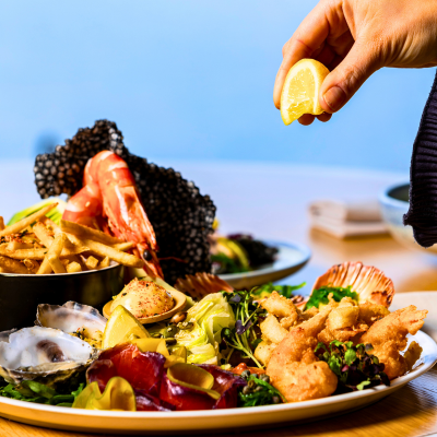 A plate of assorted seafood featuring shrimp, oysters, fried calamari, and mixed greens, with a hand squeezing a lemon wedge over the dish. Additional plates and a stack of napkins are visible on a wooden table in the background.