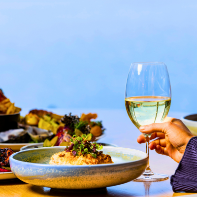 Close-up of a dining table featuring a large bowl of gourmet food garnished with greens, a hand holding a glass of white wine, and various dishes including fries and seafood in the background, suggesting an elegant restaurant setting.