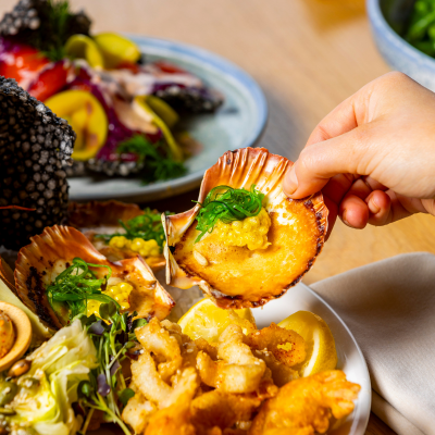 Close-up of a hand holding a scallop shell filled with seafood, garnished with greens and sauce, above a plate containing assorted seafood including fried items, lemon wedges, and greens, with another plate of food in the background.