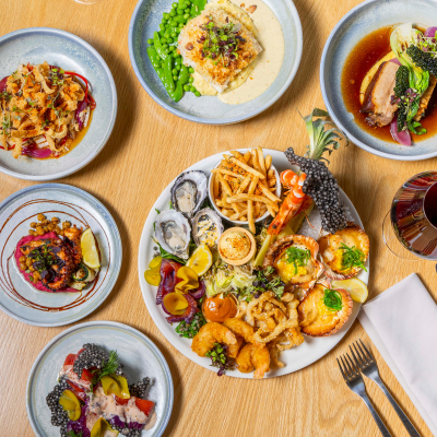 A wooden table set with a variety of plated dishes, including a central plate of seafood with oysters, prawns, calamari, and fries. Surrounding it are plates featuring fish with peas in creamy sauce, duck breast with vegetables, crab cakes with lime wedges, a salad with caviar-like garnish, and a vegetable dish with tomatoes and greens. A glass of red wine and cutlery are also visible.