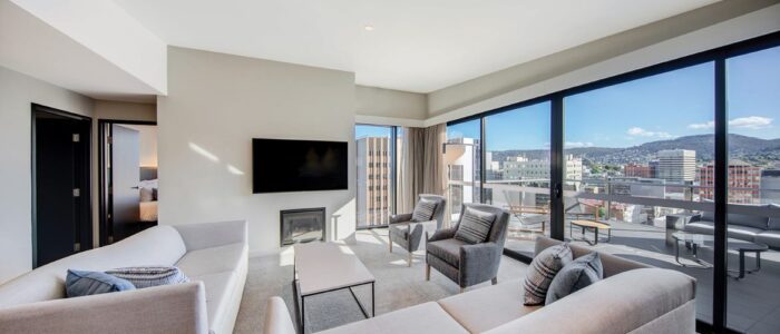 A modern living room with large floor-to-ceiling windows offering a panoramic view of a cityscape and distant mountains. The room features light gray sofas, armchairs, a coffee table, and a flat-screen TV mounted above a small fireplace. An open doorway on the left leads to another room.