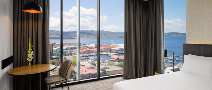 A modern hotel room with a large window offering a panoramic view of Hobart. The room includes a neatly made bed with white linens, an orange bench at the foot of the bed, and a small round table with two chairs—one patterned and one orange. A black pendant lamp hangs above the table, and long gray curtains frame the window. Outside, buildings with red roofs, water bodies, boats at piers, and distant hills are visible under a partly cloudy sky.