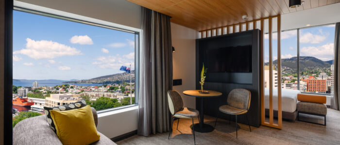 A modern hotel room with large windows showcasing a scenic view of a cityscape and distant hills. The room includes a wooden ceiling, grey carpeted floor, and contemporary furniture: a sofa with yellow and patterned cushions, a small round table with two chairs, and a partially visible bed. A flat-screen TV is mounted on the wall opposite the table.