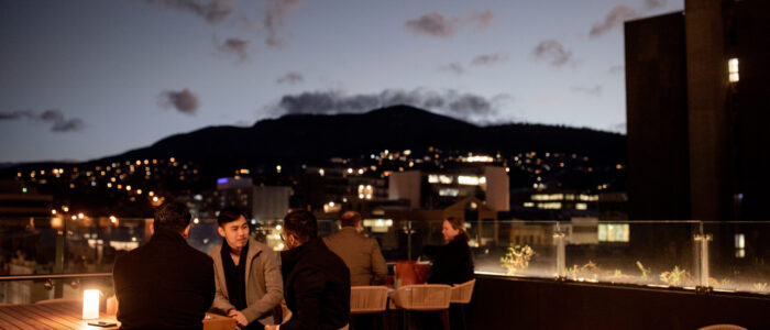 An outdoor rooftop seating area at dusk or early evening, with several people sitting at tables and conversing. The background includes a cityscape with buildings and a mountain range, illuminated by lights. The sky is darkening with visible clouds.