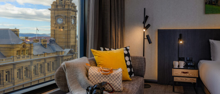 A cozy hotel room with a large window offering a view of a historic building with a clock tower and part of the cityscape. Inside, a chaise lounge with pillows, a blanket, a camera, and a bag is visible. The room also includes part of a bed, bedside tables, and modern lighting fixtures.