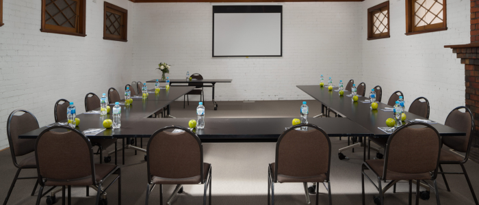 Conference room with a U-shaped table arrangement, each seat set with a bottle of water, an apple, and papers. A projector screen is mounted on the far wall, and the room features white brick walls and wooden-framed windows. A small table at the back holds flowers and other items.