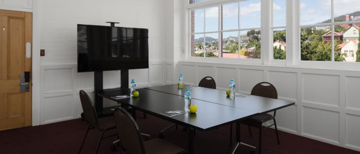 Small conference room with a rectangular table surrounded by four chairs. The table holds four water bottles and three green apples. A large flat-screen TV is mounted on a stand at one end. The room features white walls with wainscoting and large windows showing buildings and greenery outside.