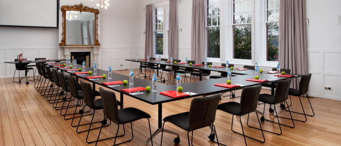 Elegant conference room with two long black tables in a U-shape, each seat set with a red folder, green apple, and water bottle. The room features large windows with gray curtains, white paneled walls, wooden flooring, a fireplace with an ornate mirror above it, and modern ceiling lights.
