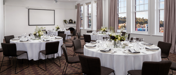 Decorated dining room set for an event with four round tables covered in white tablecloths, floral centerpieces, and full place settings. Brown chairs surround each table. The room features large windows with beige curtains, a blank projector screen, and a fireplace adorned with plants.