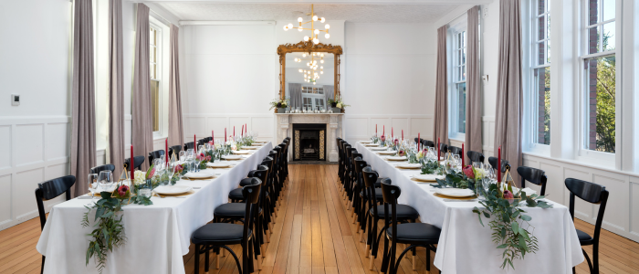 Elegant dining room set for an event with two long tables covered in white tablecloths, decorated with red candles, greenery, and full place settings. Black chairs line the tables. The room features wooden floors, white wainscoted walls, large windows, a fireplace with an ornate mirror, and ceiling lights.