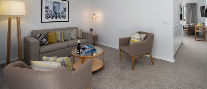 Modern living room with a grey sofa, two beige armchairs, and a round wooden coffee table holding a bottle of wine, two glasses, and books. A floor lamp with a beige shade stands beside the sofa, which has artwork above it. A small side table with decor is nearby. The room features light grey carpet, white walls, and an open doorway leading to another room.