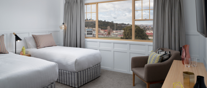 Neatly arranged hotel room with two single beds featuring white bedding and striped pillows. A large window with grey curtains offers a view of trees and buildings. The room includes a cushioned armchair, a wooden desk or dresser with decorative vases and glasses, and wall-mounted lamps above the beds.